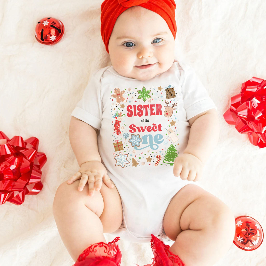 Smiling baby girl in a red headband wearing a white Sister of the Sweet One bodysuit among red bows.