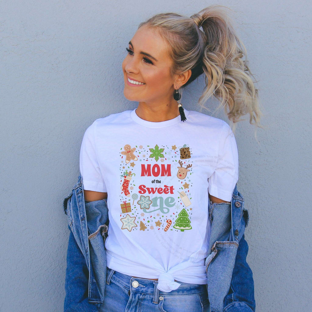Smiling woman in a denim jacket wearing a white Mom of the Sweet One shirt covered in Christmas cookies.