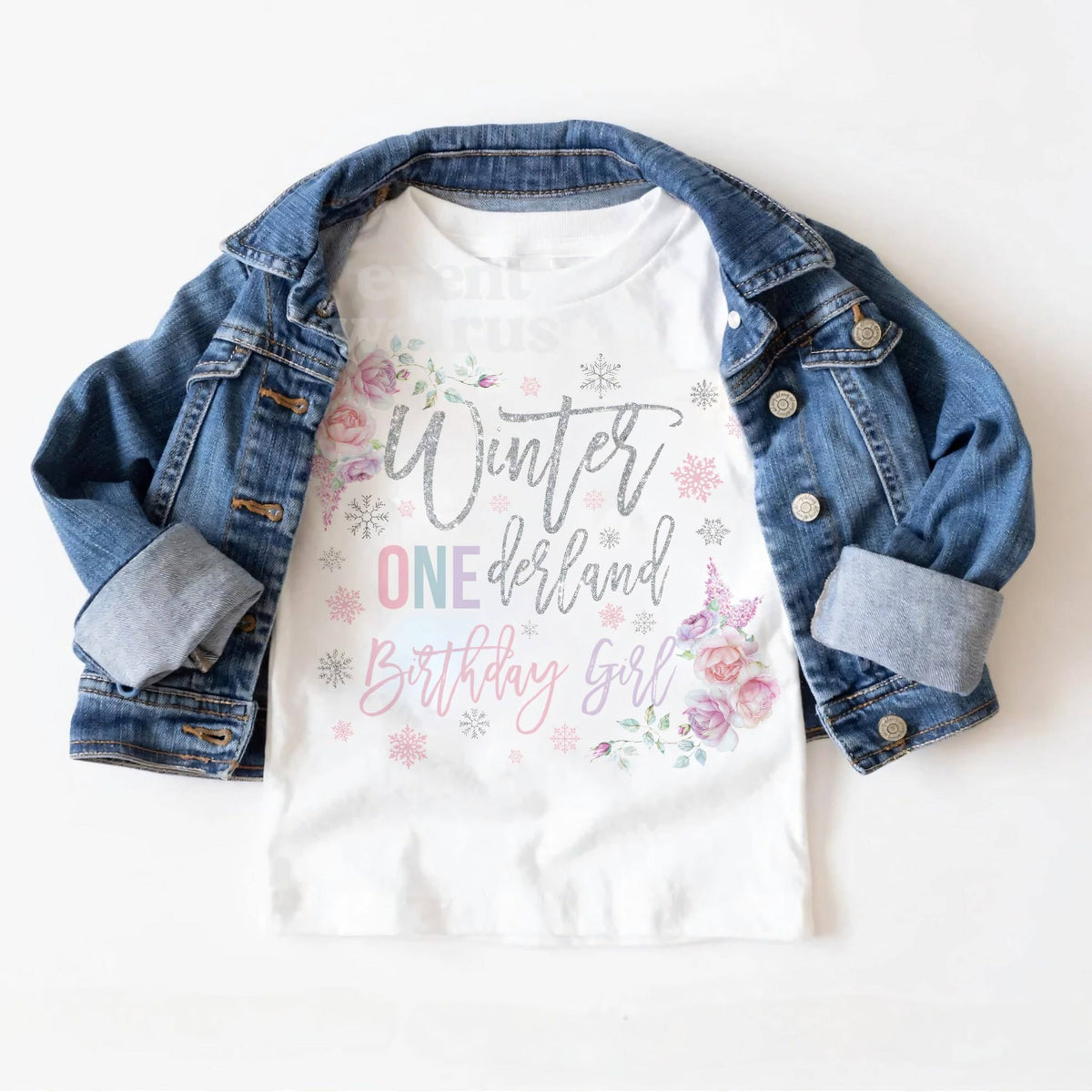 White floral Winter ONEderland Birthday Girl toddler tee laid on a denim jacket.
