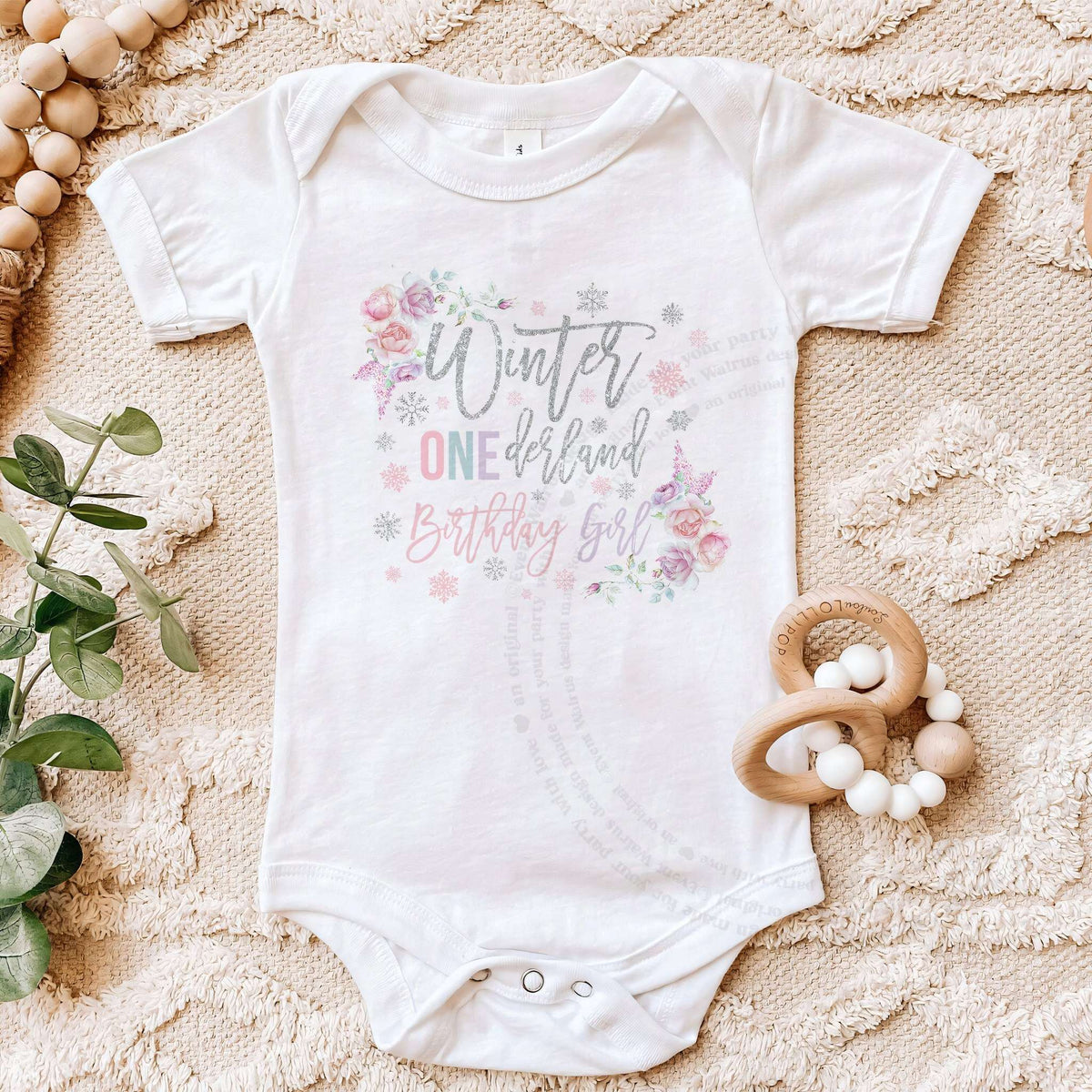 White floral Winter ONEderland Birthday Girl bodysuit on a beige blanket with a wooden teether.