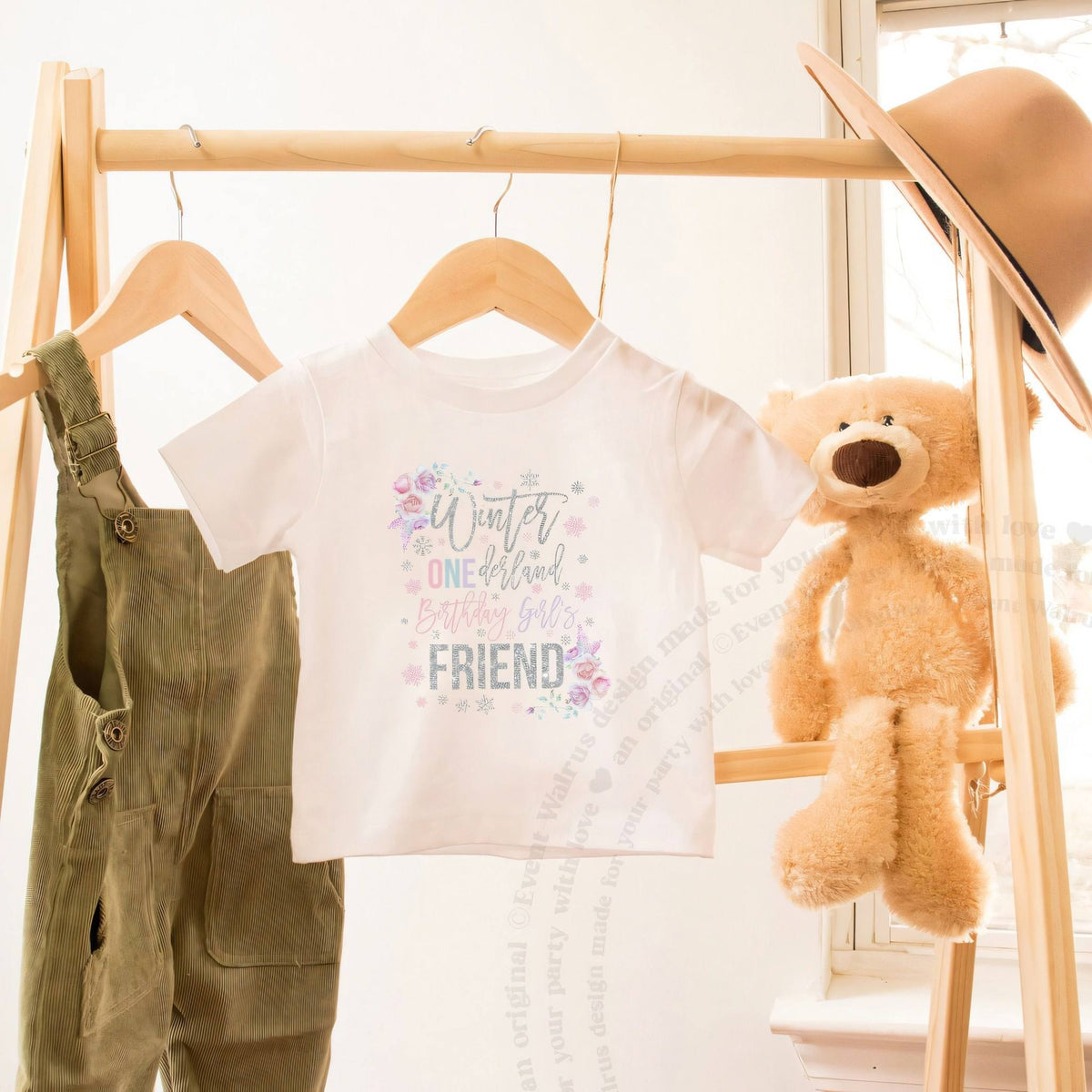 White floral Winter ONEderland Friend toddler tee on a wooden clothes rack beside a teddy bear.