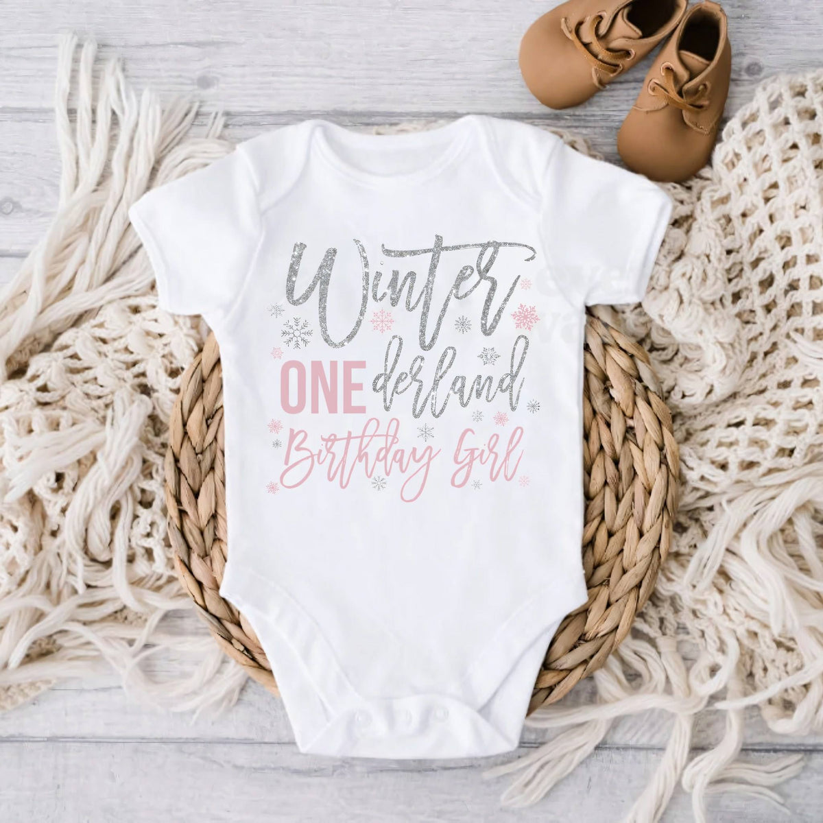 White Winter ONEderland Birthday Girl bodysuit on gray wood with a knit blanket and baby shoes.