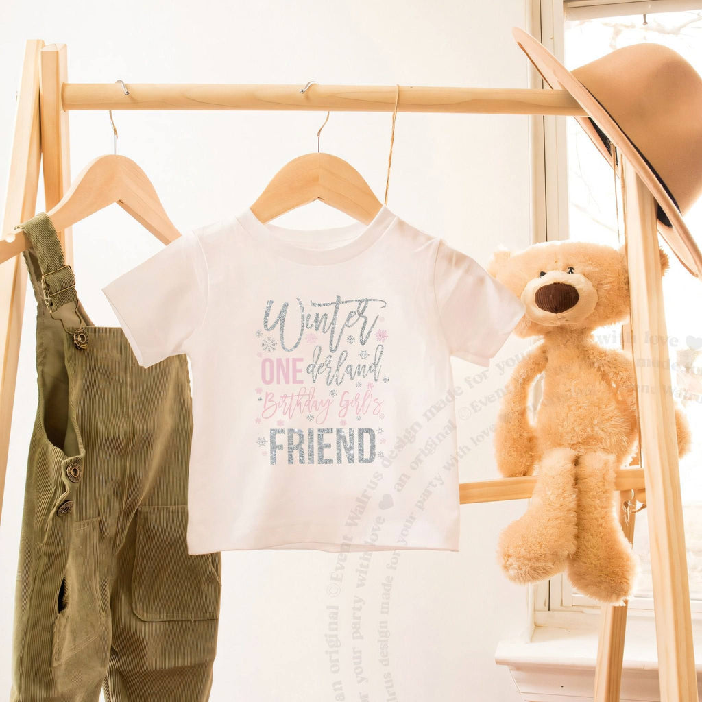 White Winter ONEderland Friend toddler tee on a wooden clothes rack beside a teddy bear.