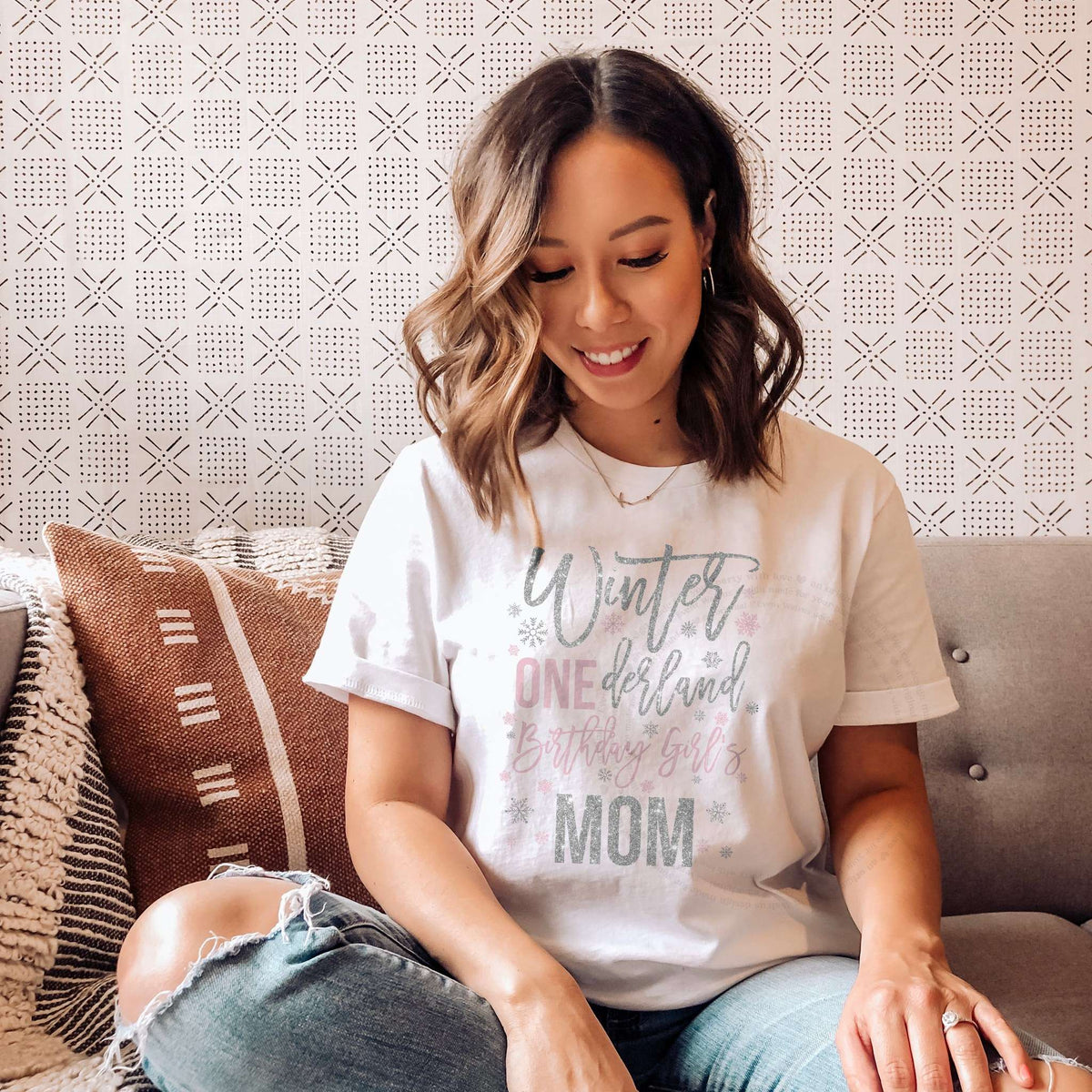 Woman sitting on a sofa with patterned pillows, wearing a white Winter Onederland Mom shirt in pink and silver glitter.