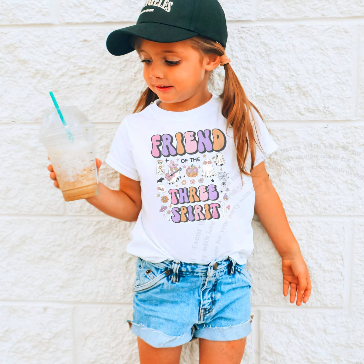 Girl in a ball cap holding a drink in a white Friend of the Three Spirit shirt.