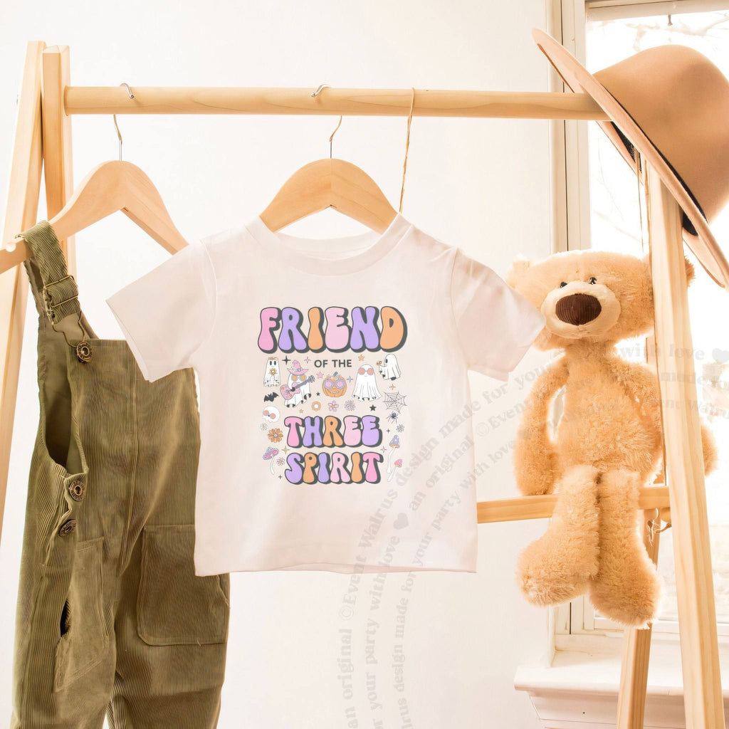 White Friend of the Three Spirit toddler tee on a wooden clothes rack beside a teddy bear.