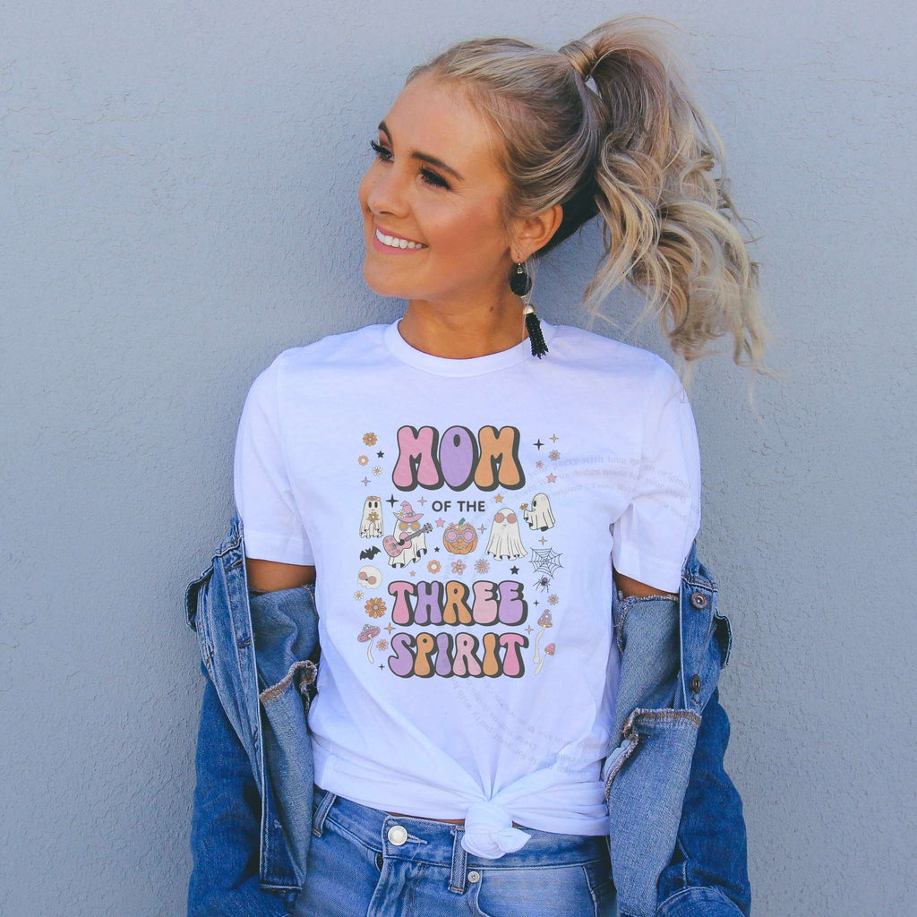 Woman in a denim jacket wearing a Mom of the Three Spirit shirt with groovy pastel ghosts.