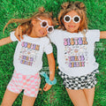 Two girls in heart sunglasses lying on grass in white Sister and Cousin Three Spirit shirts.