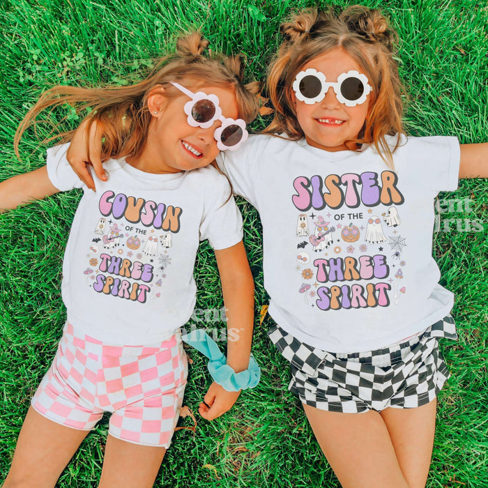 Two girls in heart sunglasses lying on grass in white Sister and Cousin Three Spirit shirts.