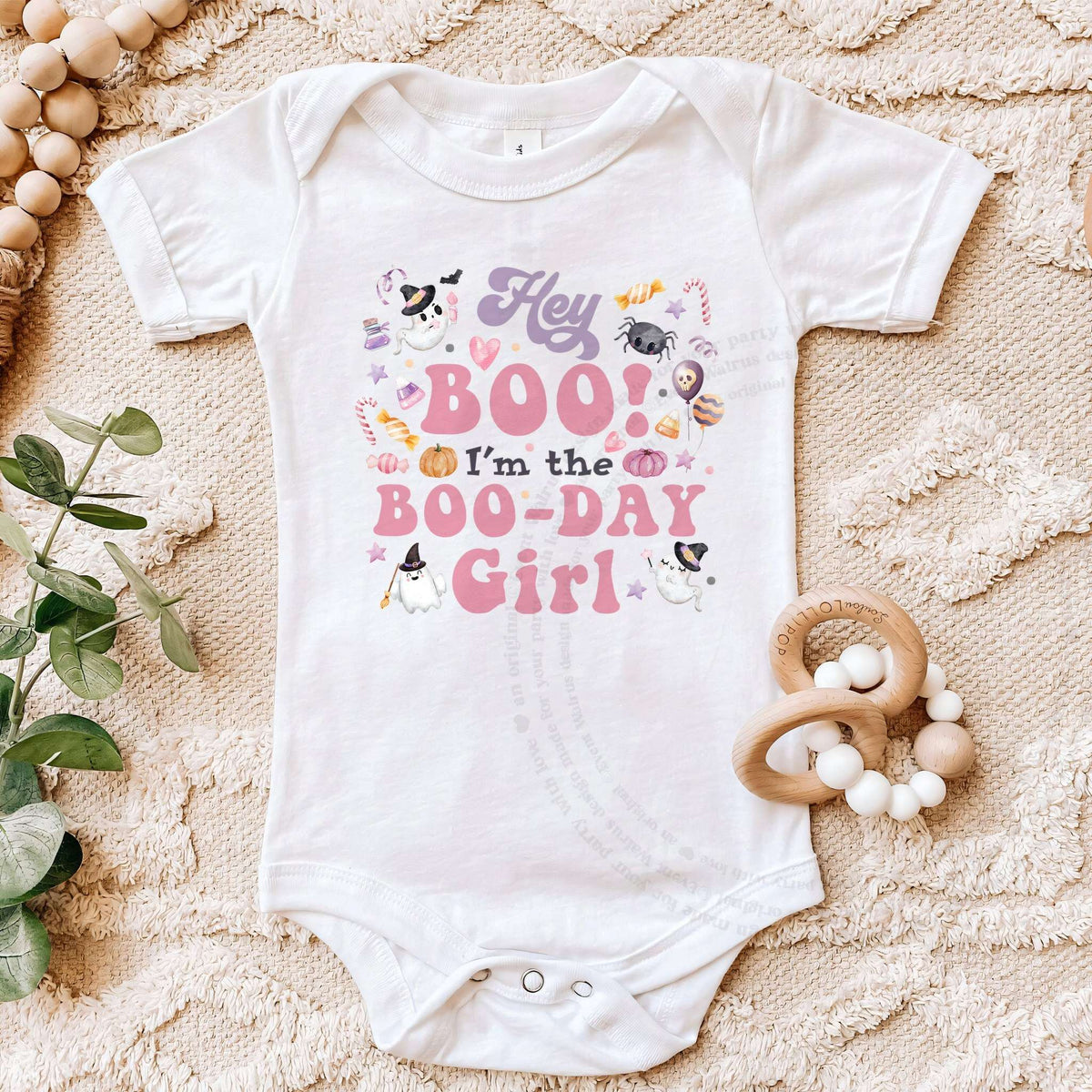 White Hey Boo Boo-Day Girl bodysuit on a beige blanket with a wooden teether.