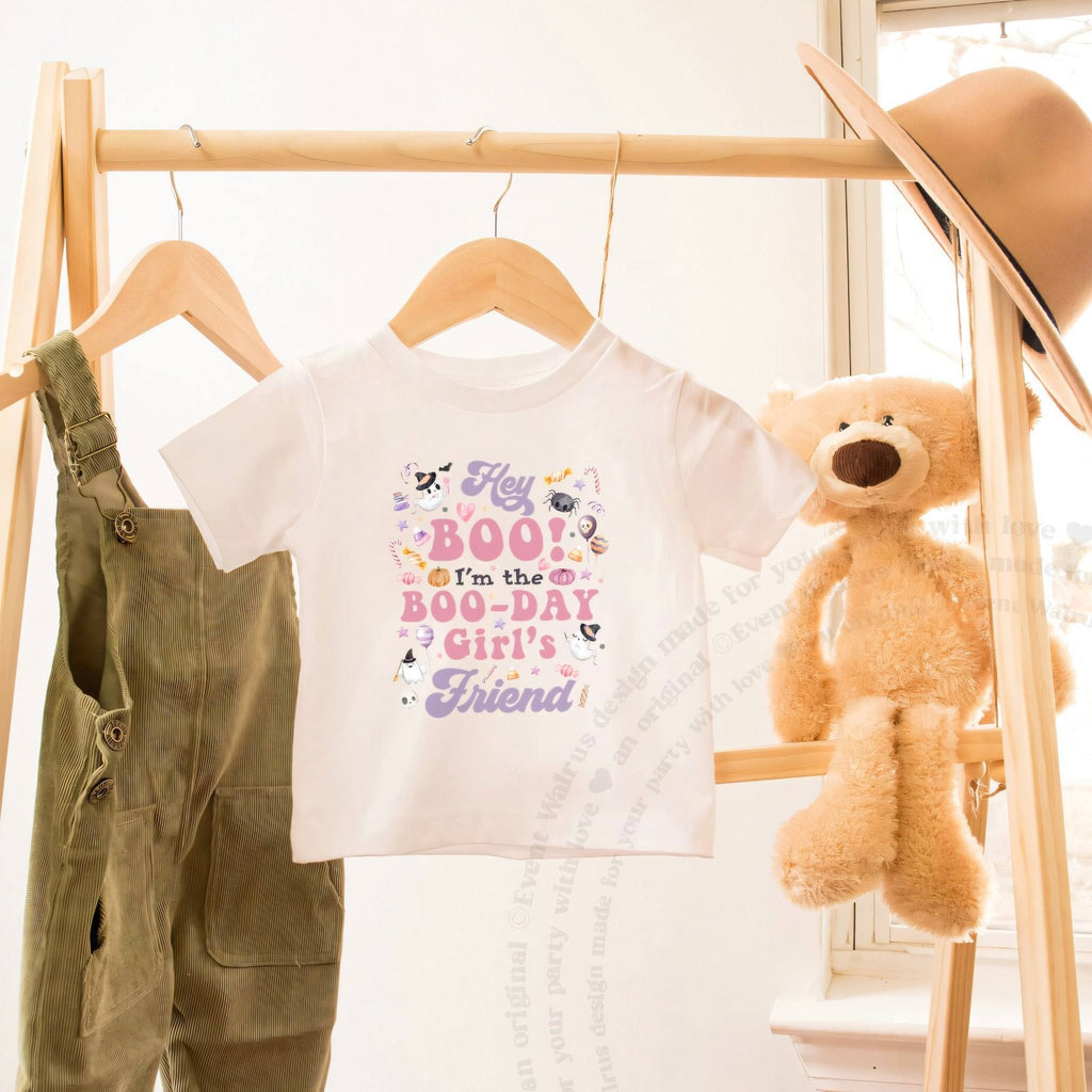 White Hey Boo Friend toddler tee on a wooden clothes rack beside a teddy bear.