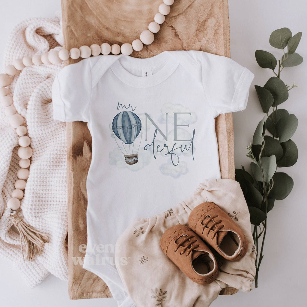 White Mr Onederful bodysuit laid flat with linen bloomers and a wooden bead garland.