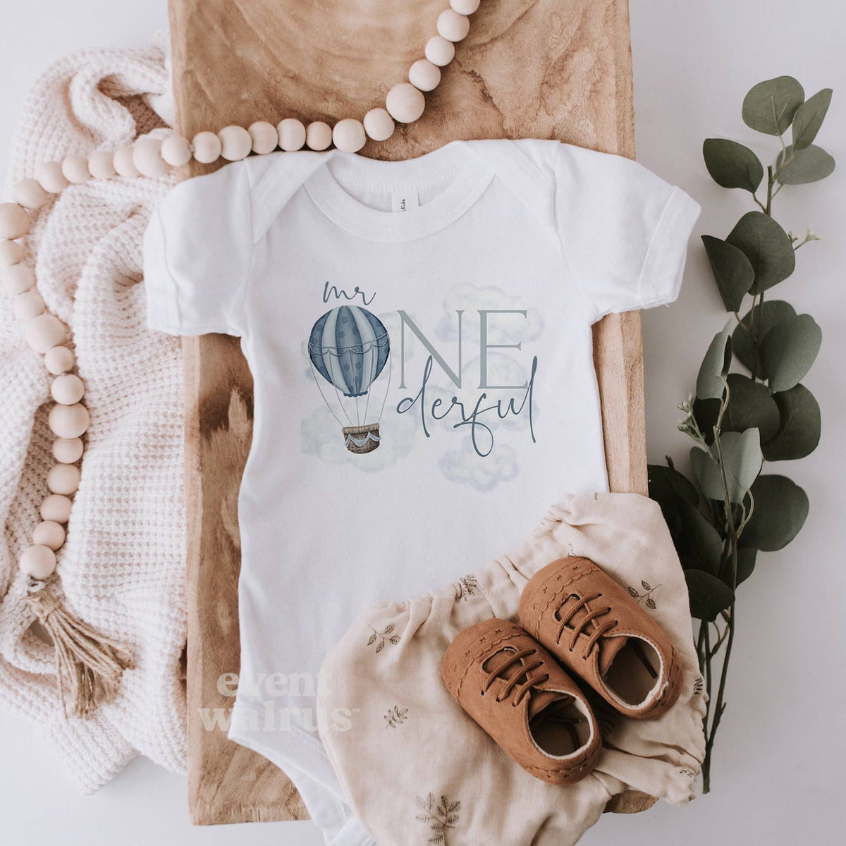 White Mr Onederful bodysuit laid flat with linen bloomers and a wooden bead garland.