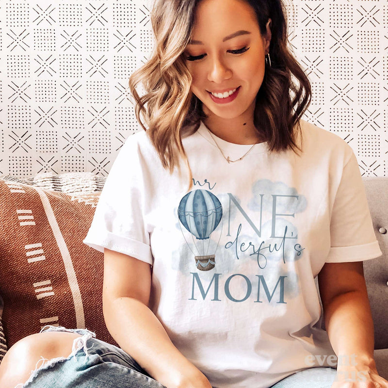 Smiling mom on a sofa in a white Mr Onederful's Mom shirt with a blue hot air balloon.