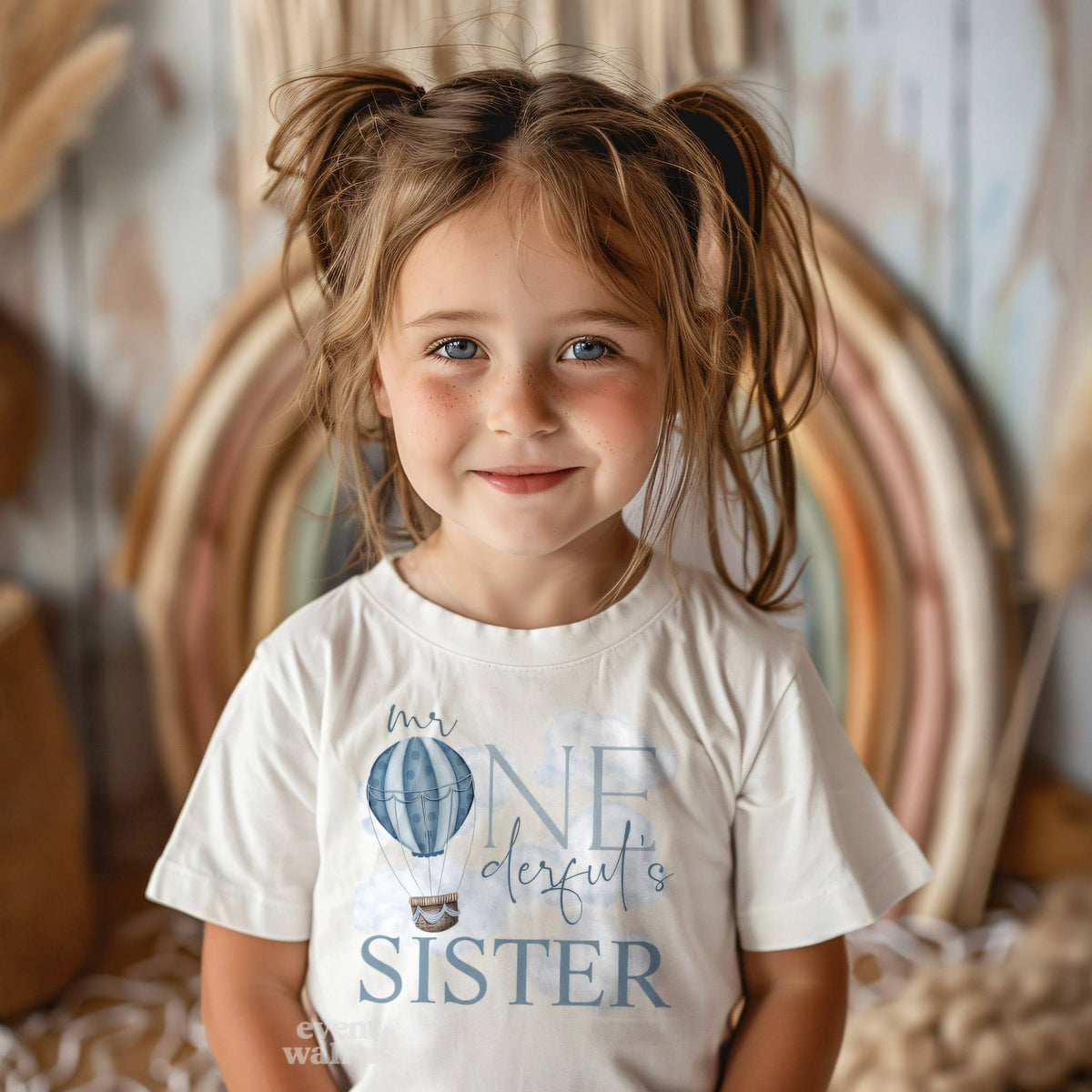 Smiling toddler girl in a white Mr Onederful's Sister tee in front of a rainbow.