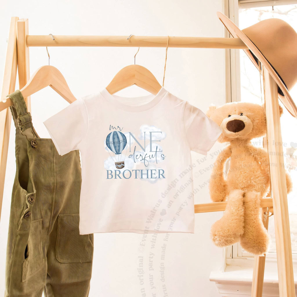 White Mr Onederful's Brother toddler tee on a wooden hanger beside a teddy bear.