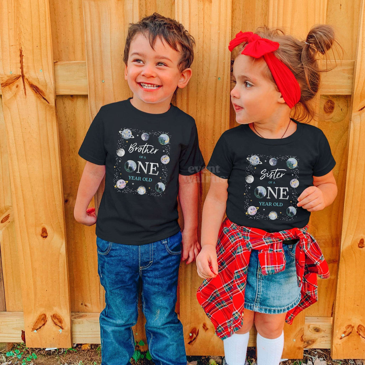 Brother and sister in black ONE Year Old space shirts by a wooden fence.