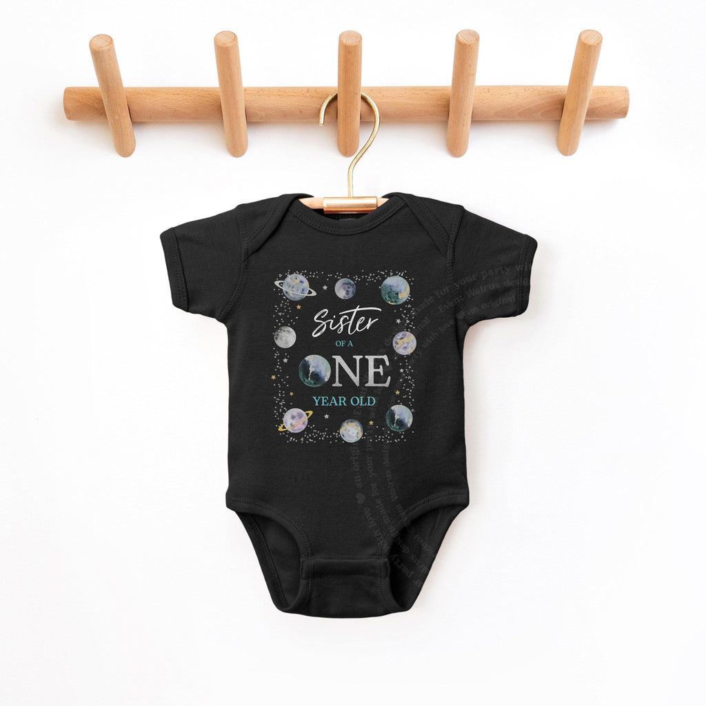 Black Sister of a ONE Year Old bodysuit hanging on a wooden peg rack.