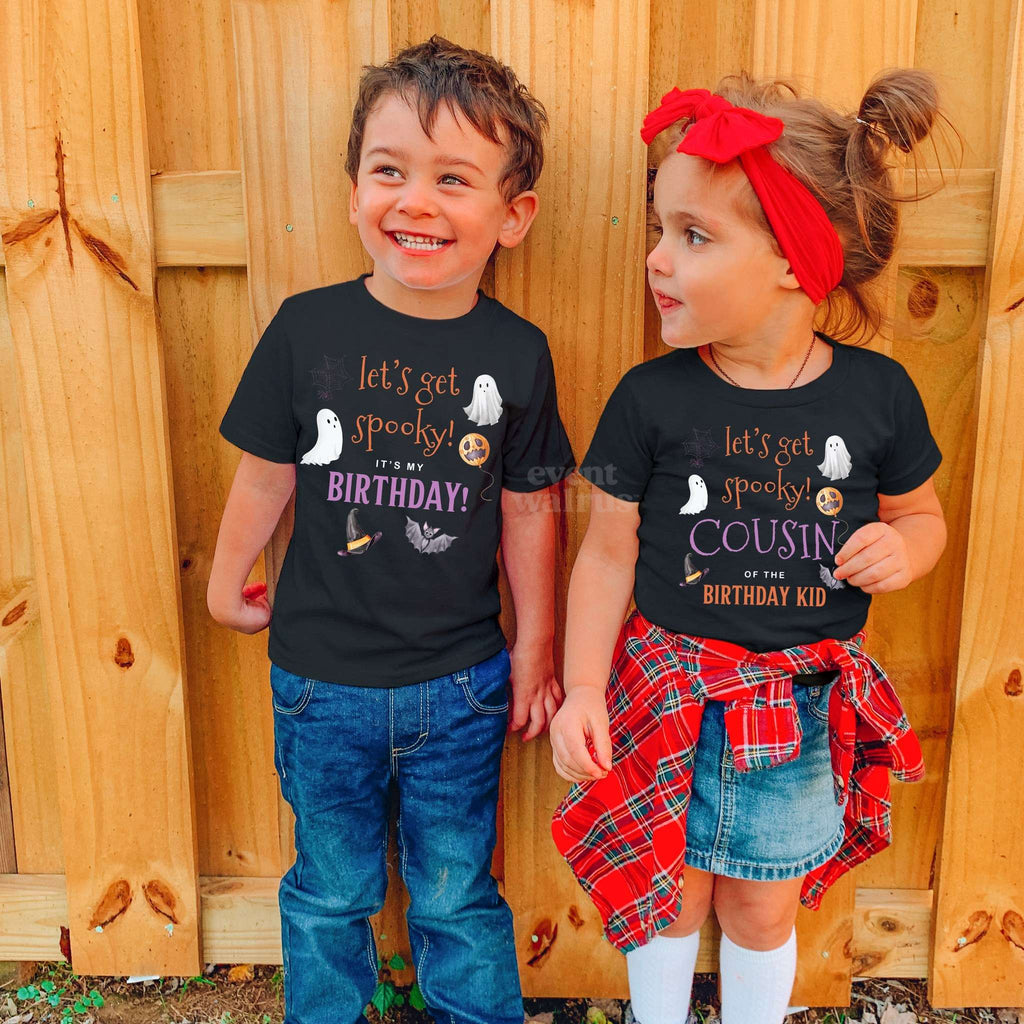 Boy and girl in black Let's Get Spooky Birthday and Cousin shirts by a wooden fence.