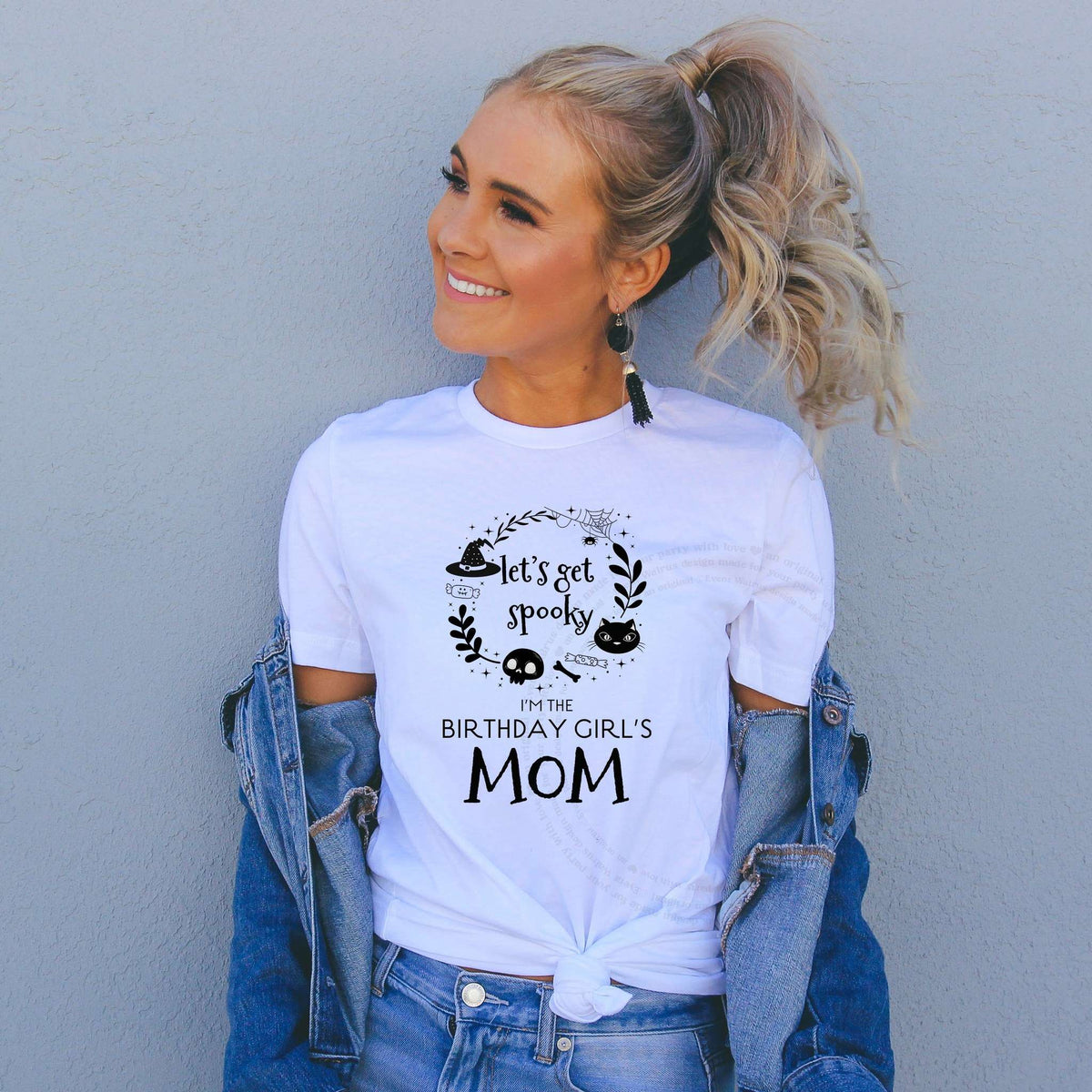 Smiling woman in a denim jacket wearing a white Let's Get Spooky Birthday Girl's Mom shirt with black doodles.