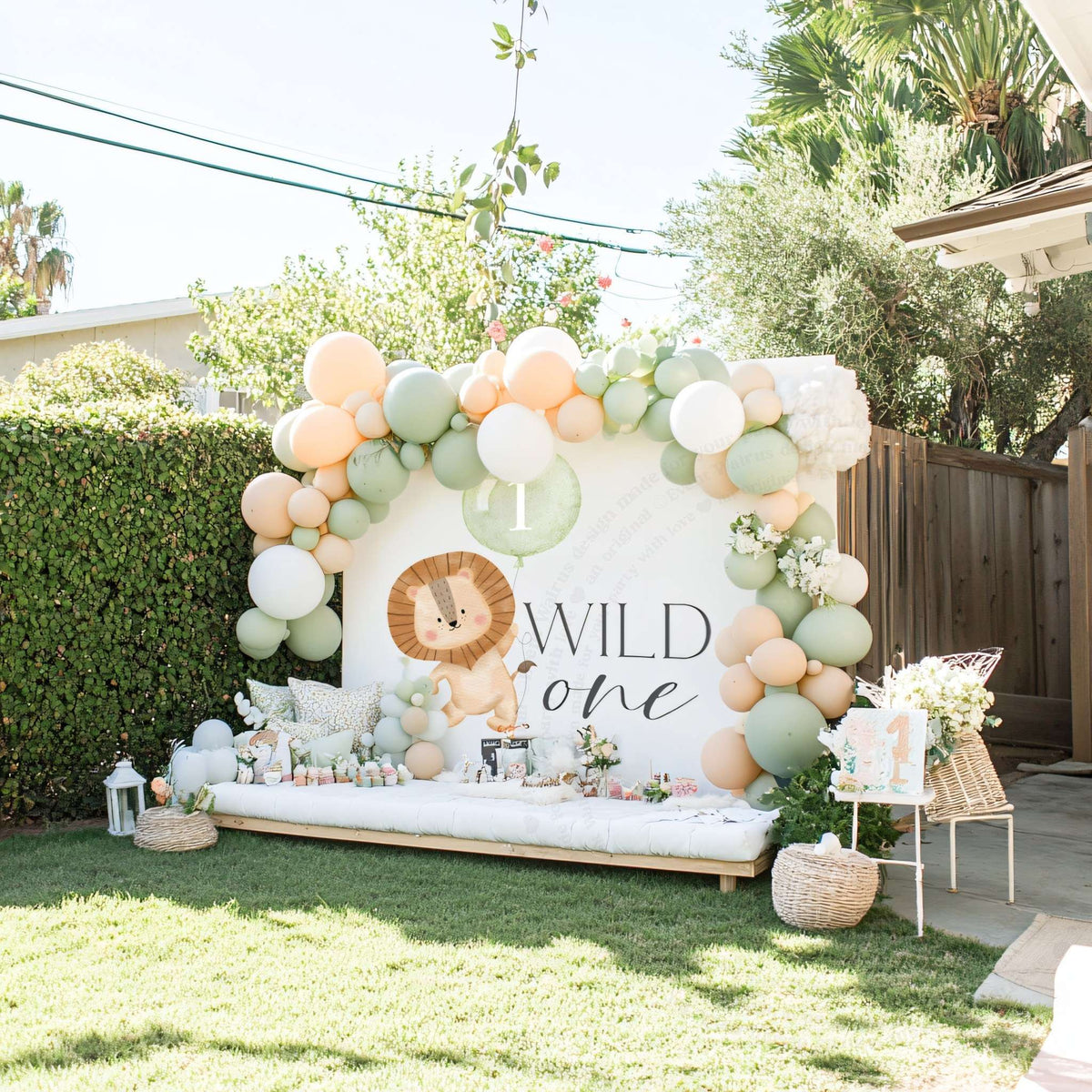 Lion sage balloon Wild One backdrop in a backyard with a peach and sage balloon arch over a low lounge.