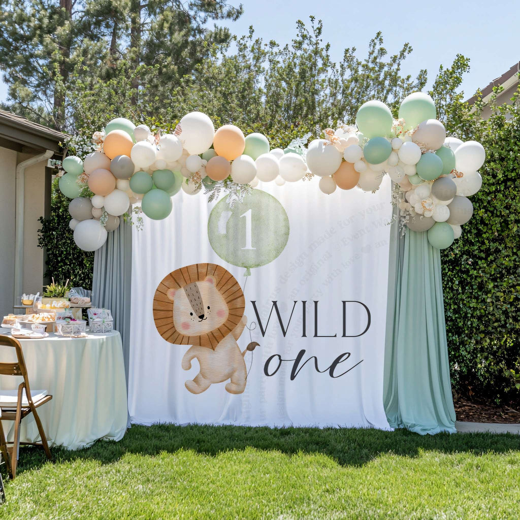 Lion first birthday backdrop between sage drapes in a garden, beside a dessert table and a balloon garland.