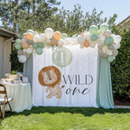 Lion first birthday backdrop between sage drapes in a garden, beside a dessert table and a balloon garland.