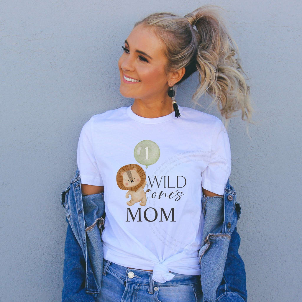Smiling woman in a denim jacket wearing a white Wild One's Mom shirt with the lion and sage green balloon.