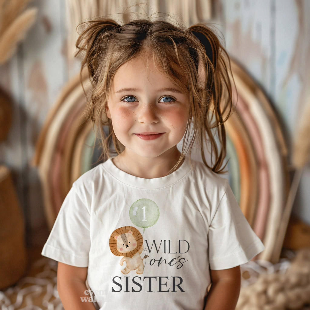 Smiling toddler girl in a white Wild One's Sister tee in front of a boho rainbow.