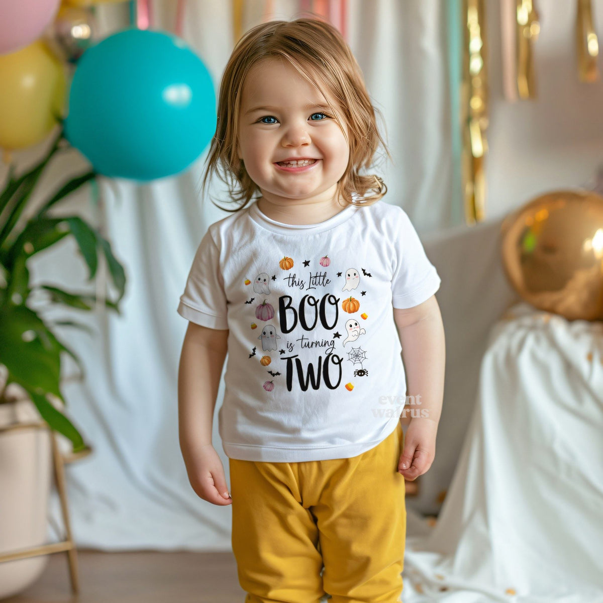 Smiling toddler girl among party balloons in a white This Little Boo Is Turning Two tee.