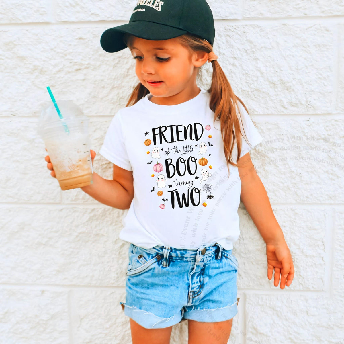 Girl in a ball cap holding a drink in a white Friend of the Little Boo shirt.