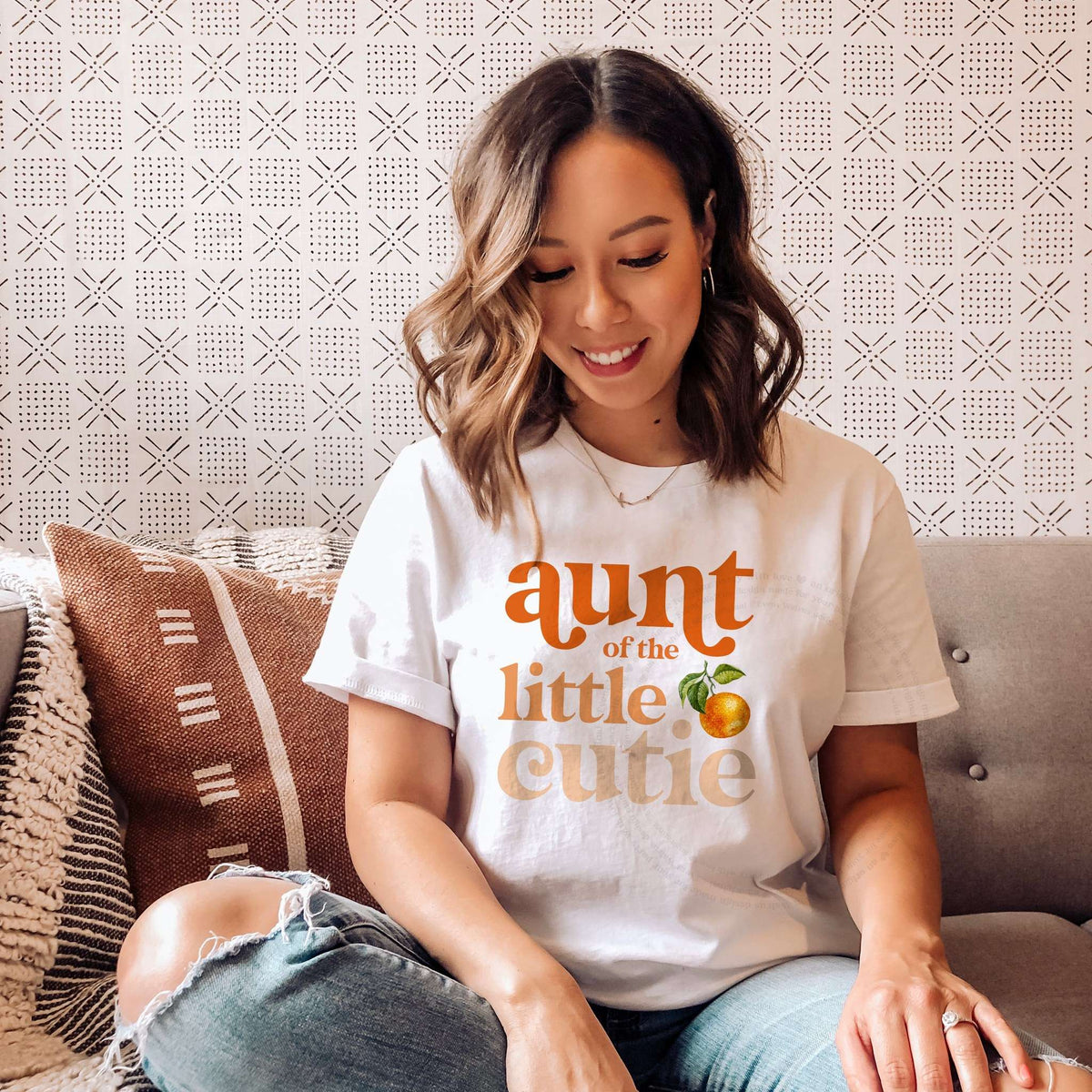 Woman sitting on a sofa in a white Aunt of the Little Cutie shirt with peach and orange letters.