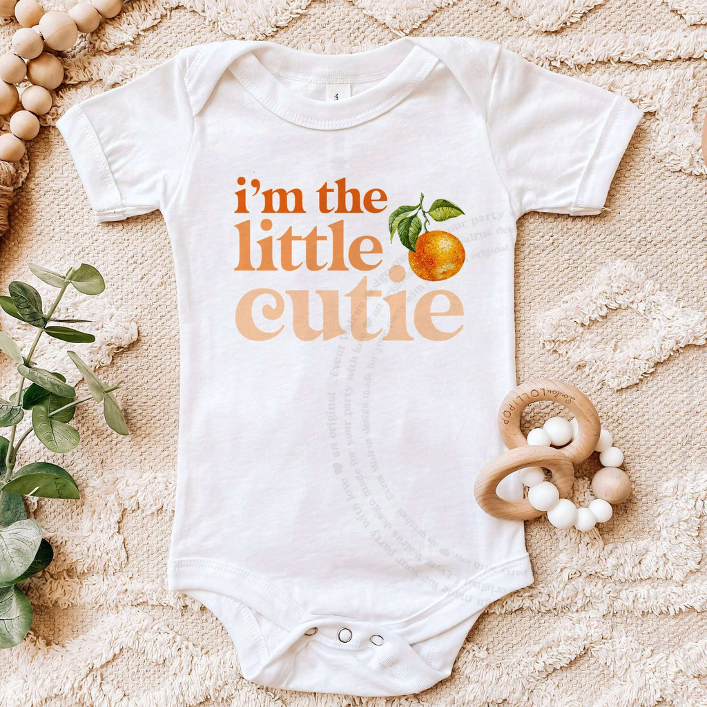 White I'm the Little Cutie bodysuit laid flat with a wooden teether and eucalyptus.