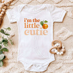 White I'm the Little Cutie bodysuit laid flat with a wooden teether and eucalyptus.