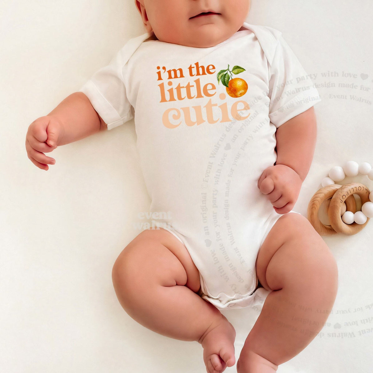 Baby lying down in a white I'm the Little Cutie bodysuit beside a wooden teether.