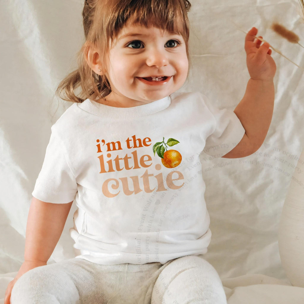 Smiling baby girl in a white I'm the Little Cutie baby tee.