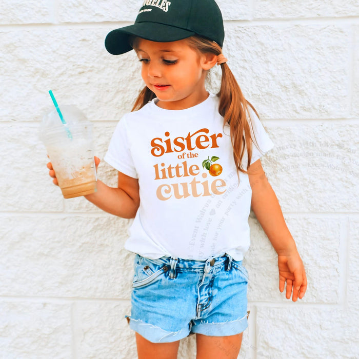 Girl in a black cap holding a drink in a white Sister of the Little Cutie tee.
