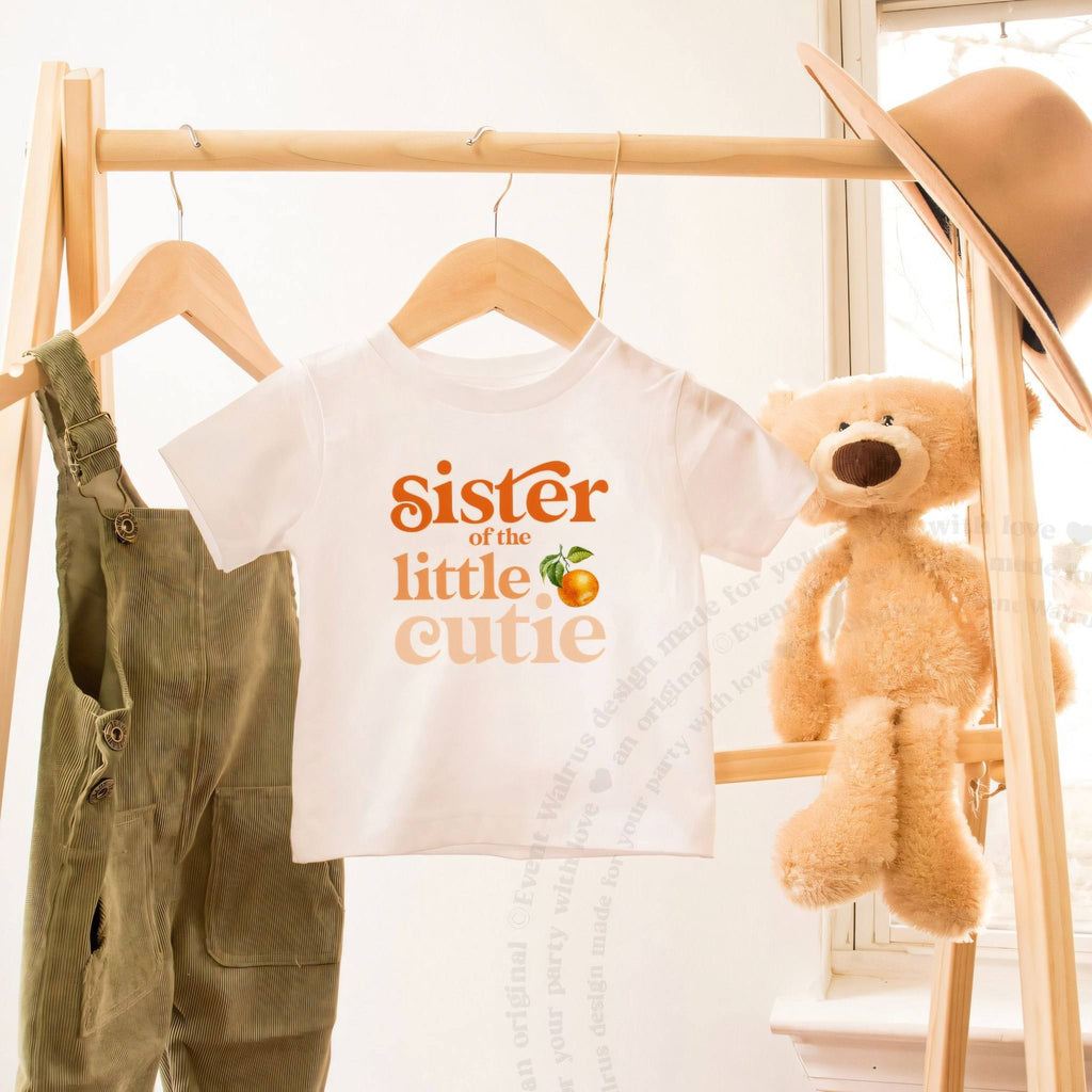 White toddler Sister of the Little Cutie tee on a wooden rack beside a teddy bear.