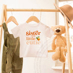 White toddler Sister of the Little Cutie tee on a wooden rack beside a teddy bear.