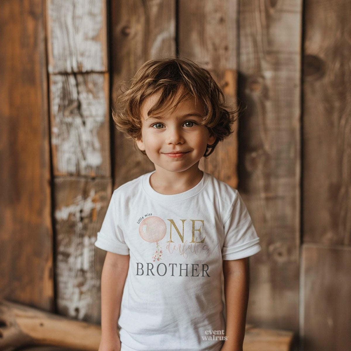 Toddler boy against a wooden wall in a white Brother shirt with a pink balloon and gold ONE.