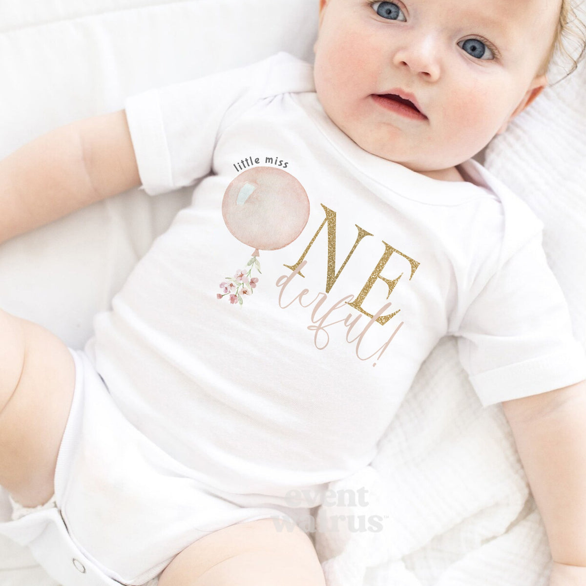 Baby girl with a pink bow lying on a white blanket in a Little Miss Onederful bodysuit.