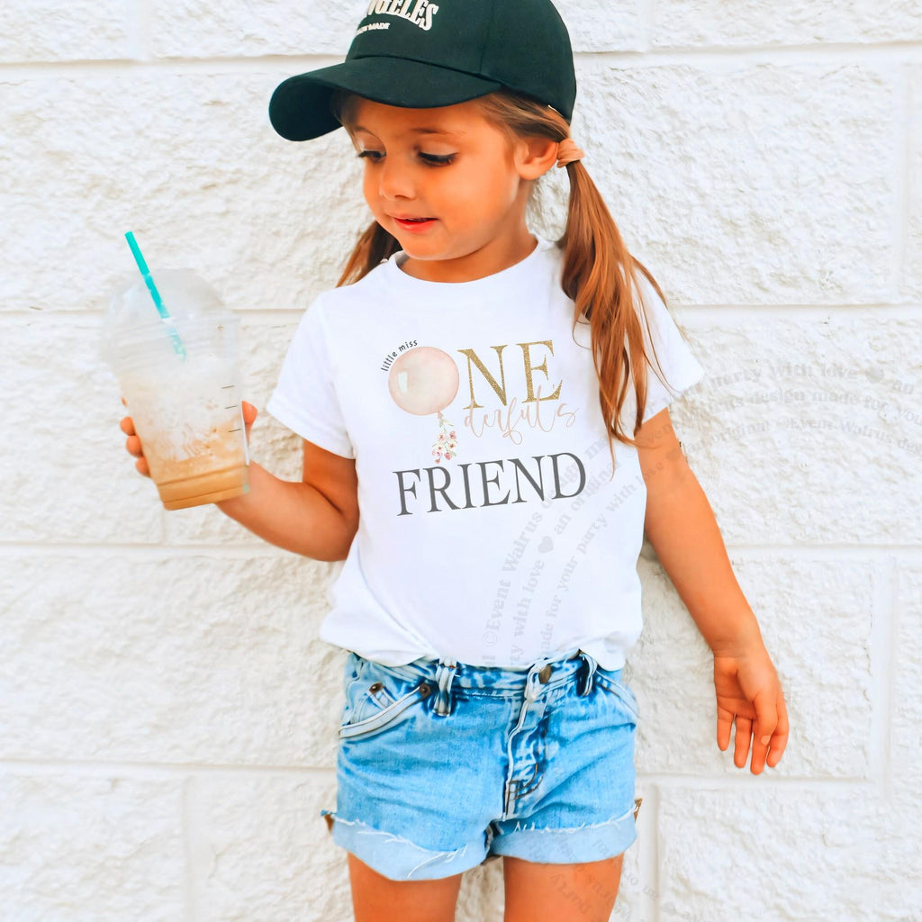 Girl in a cap holding a drink in a white Friend shirt with a pink balloon and gold ONE.