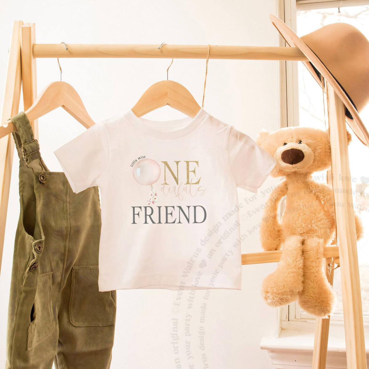 White Friend toddler tee with the pink balloon design on a wooden hanger beside a teddy bear.