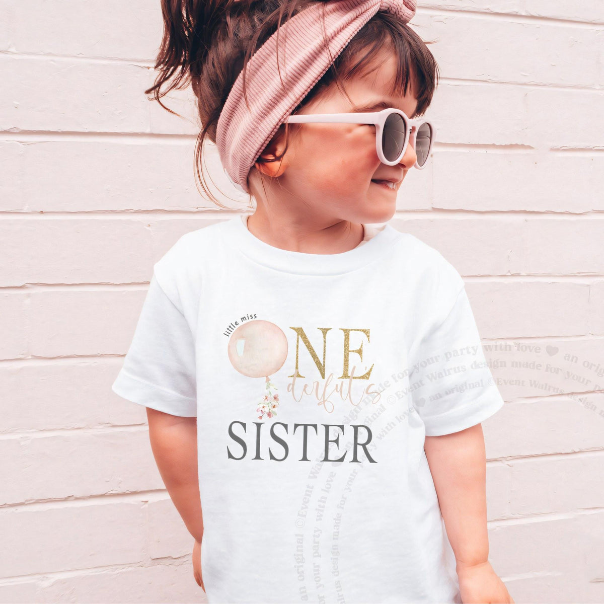 Girl in sunglasses and a headband wearing a white Sister shirt with a pink balloon and gold ONE.