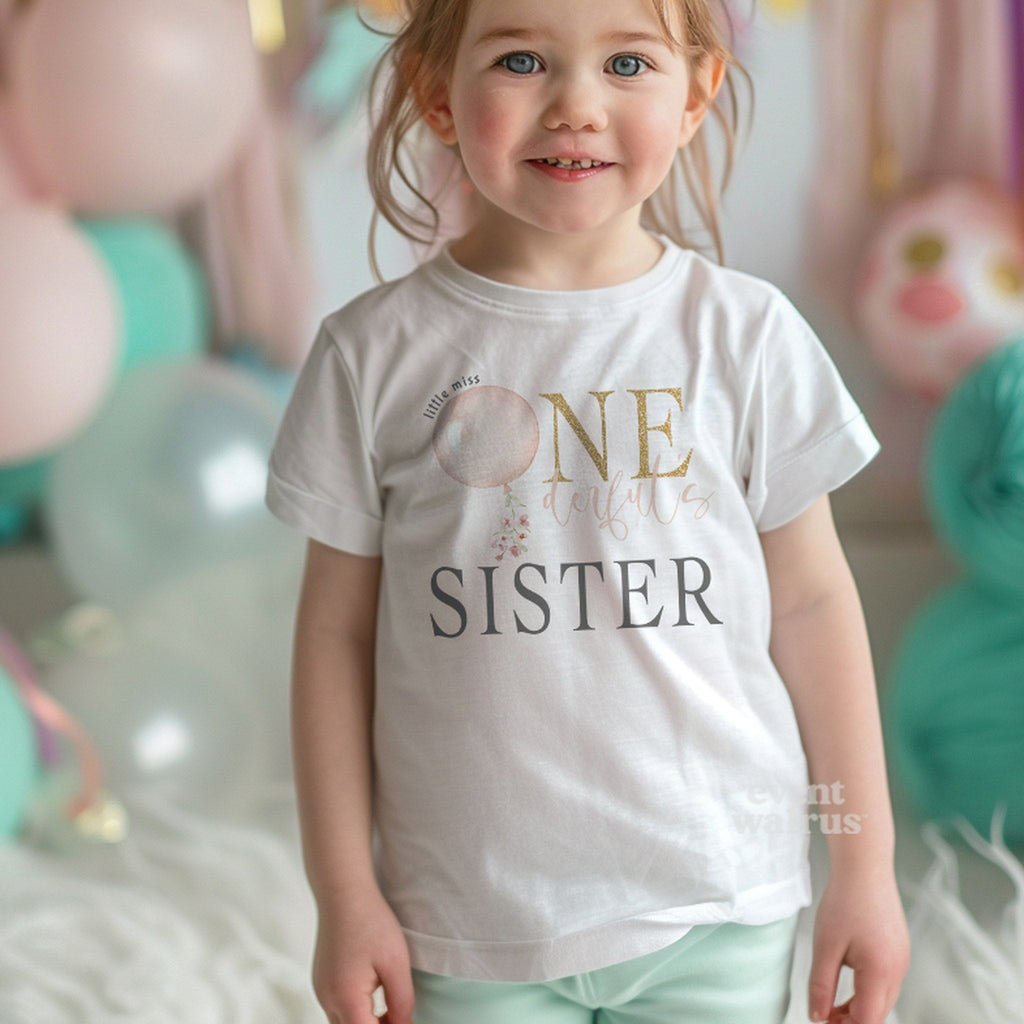 Smiling toddler girl among pastel balloons in a white Sister shirt with the pink balloon design.