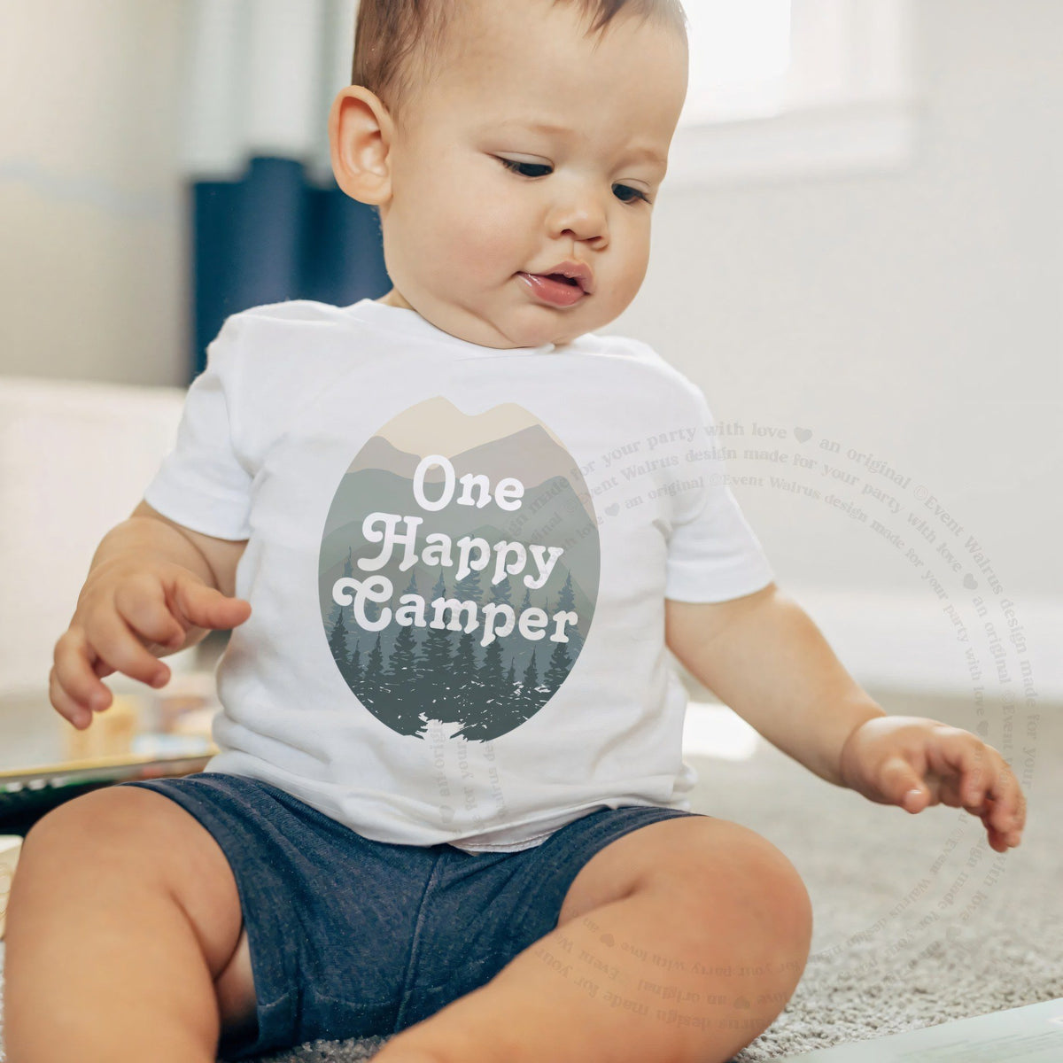 Baby boy sitting on a rug in a white One Happy Camper baby tee.