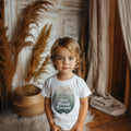 Toddler boy in a boho room wearing a white Brother of One Happy Camper tee.