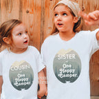 Two girls against a wood wall in white Cousin and Sister One Happy Camper shirts.