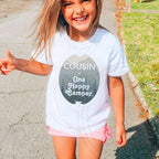 Laughing girl outdoors in a white Cousin of One Happy Camper kids tee.