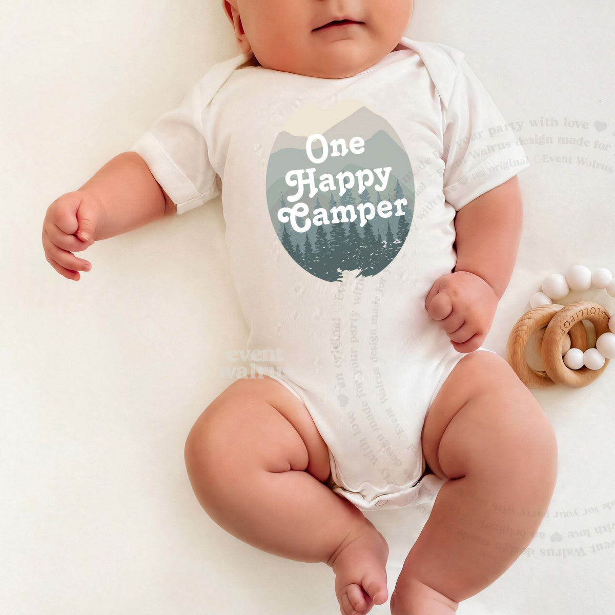 Baby lying on a white blanket in a One Happy Camper bodysuit with misty green mountains.